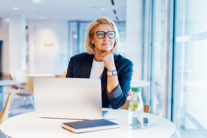 50's confident mature businesswoman dreamy looking at window, middle-aged experienced senior female professional working on laptop in open space office. Female entrepreneur working remotely.